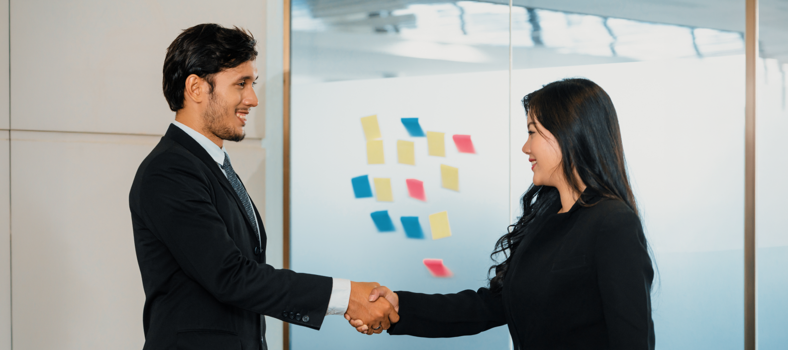 Two professionals in business attire shake hands in a bright office with colorful sticky notes on a glass wall behind them to signal a agreement or partnership.