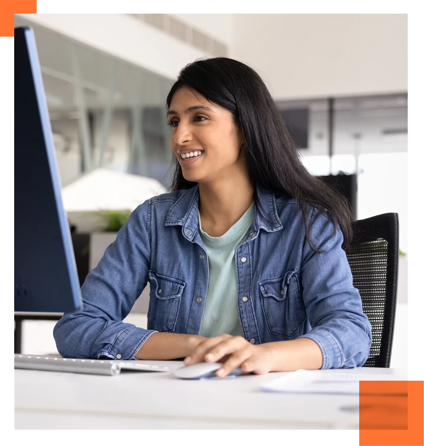 Woman smiling while working at a desktop computer in a bright office, denim jacket, light green top.