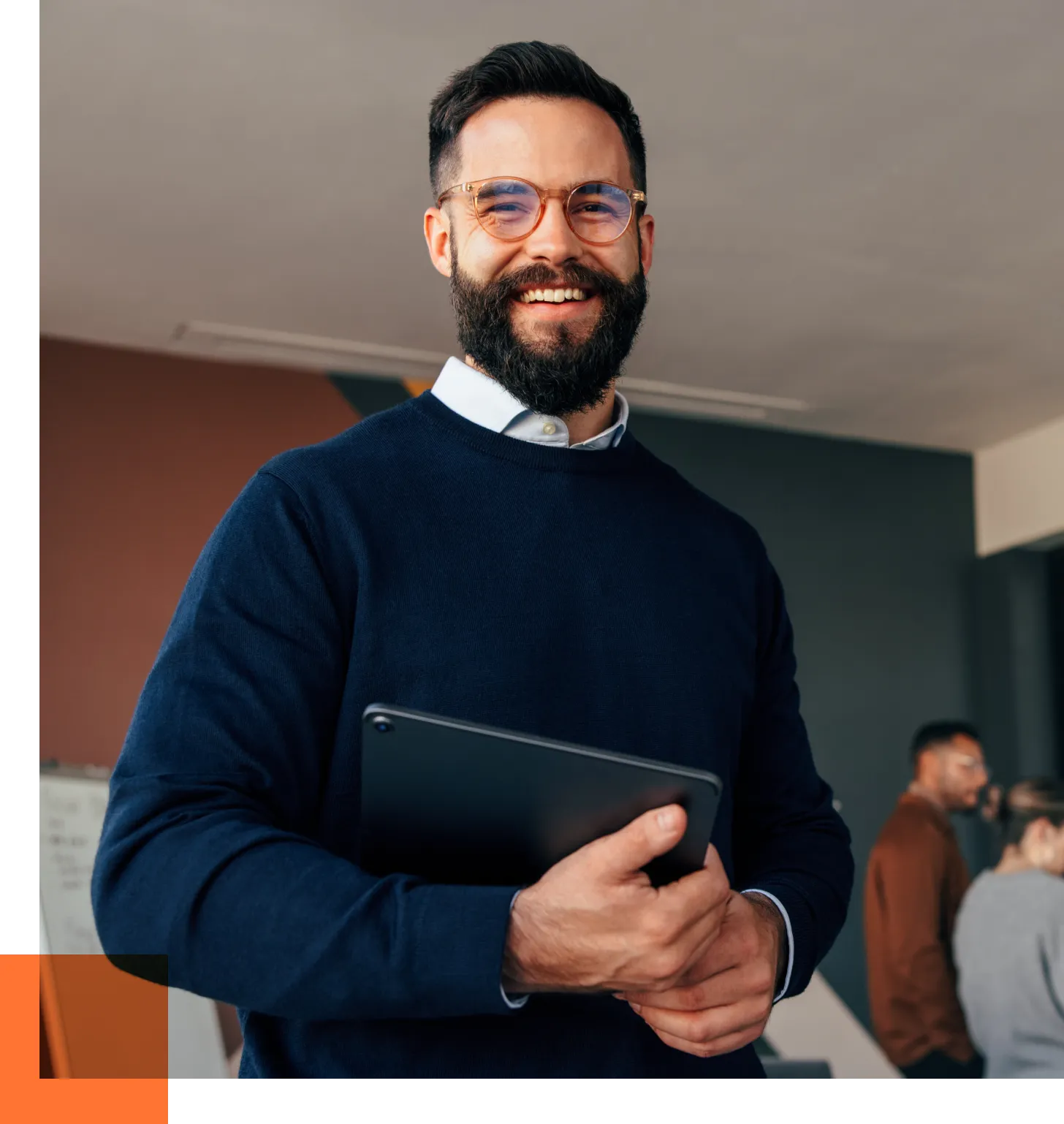 Smiling man with a beard and round glasses holding a tablet in an office setting.