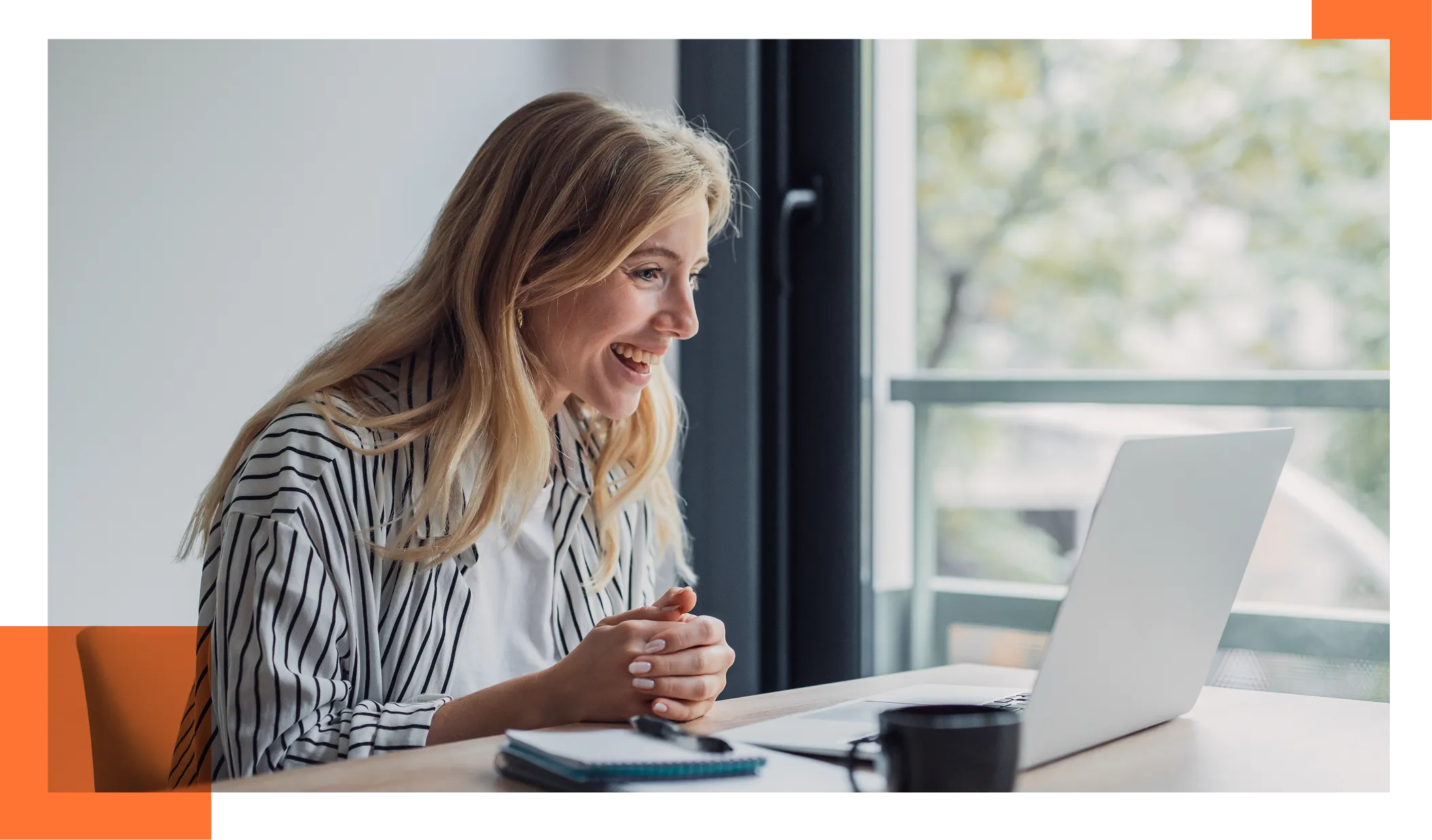 Smiling woman in a striped shirt working on a laptop at a bright desk by a window, with a notebook nearby.