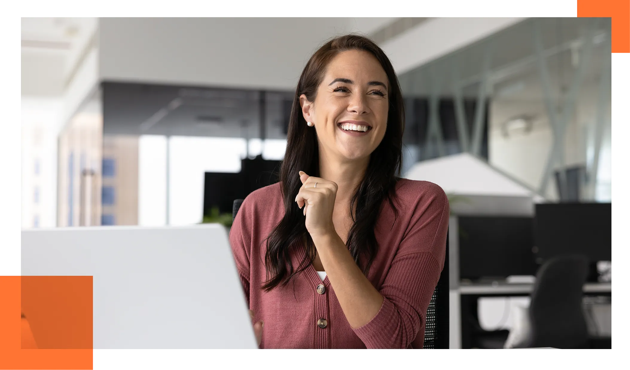 woman with long dark hair wearing a maroon cardigan smiles at her desk with a laptop in a bright, modern office.