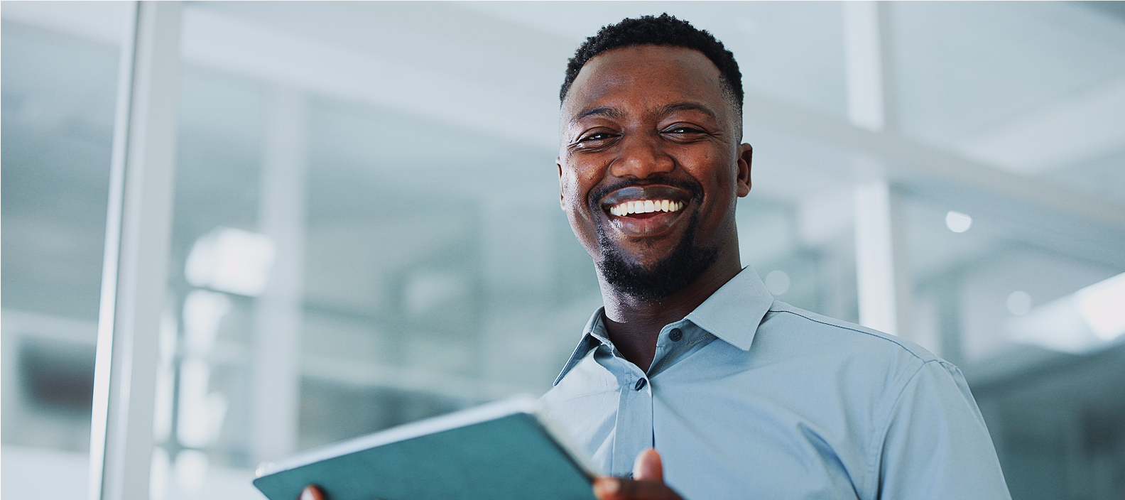 Smiling man in a light blue shirt holding a notebook in an bright office space.