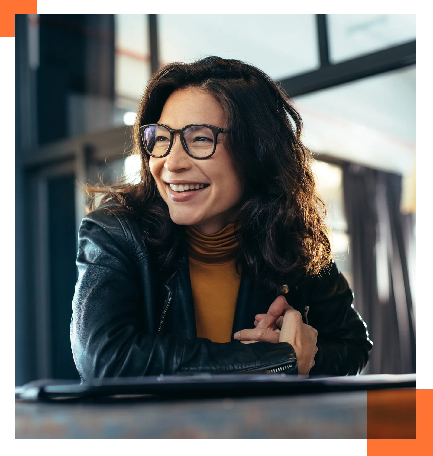 Smiling woman with black hair and glasses, wearing an orange turtleneck and black leather jacket, indoors by large windows.