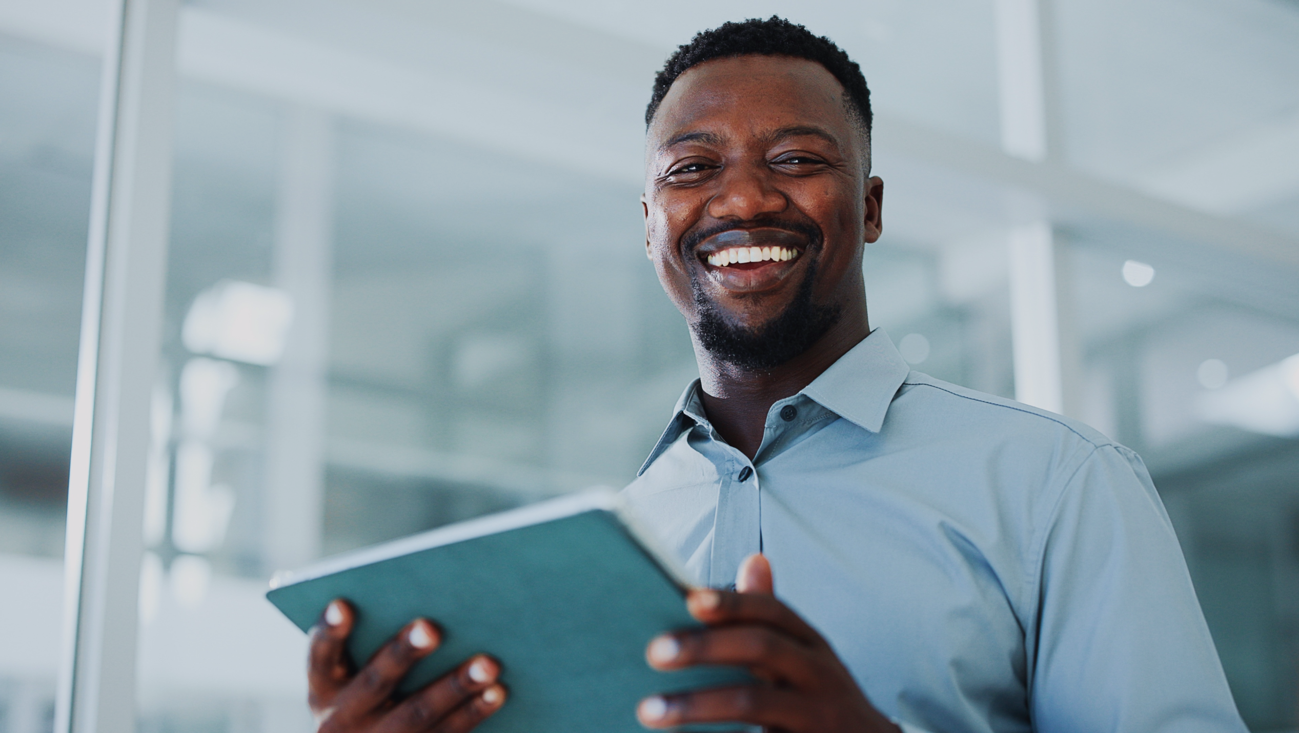 Man in a light blue shirt smiling while holding a green notebook in an office setting.