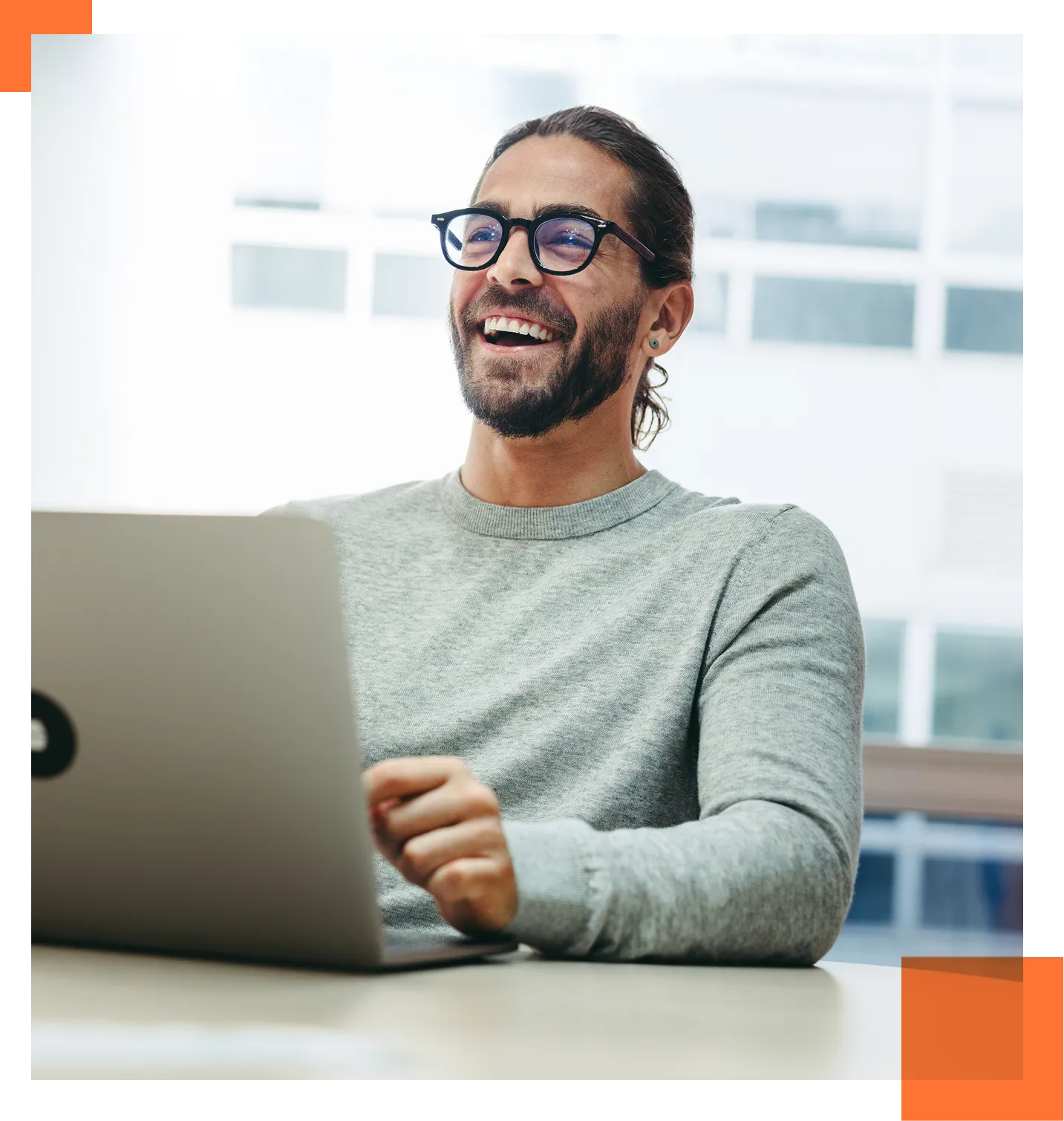 Man with glasses and beard smiling while using a laptop in a bright office environment.