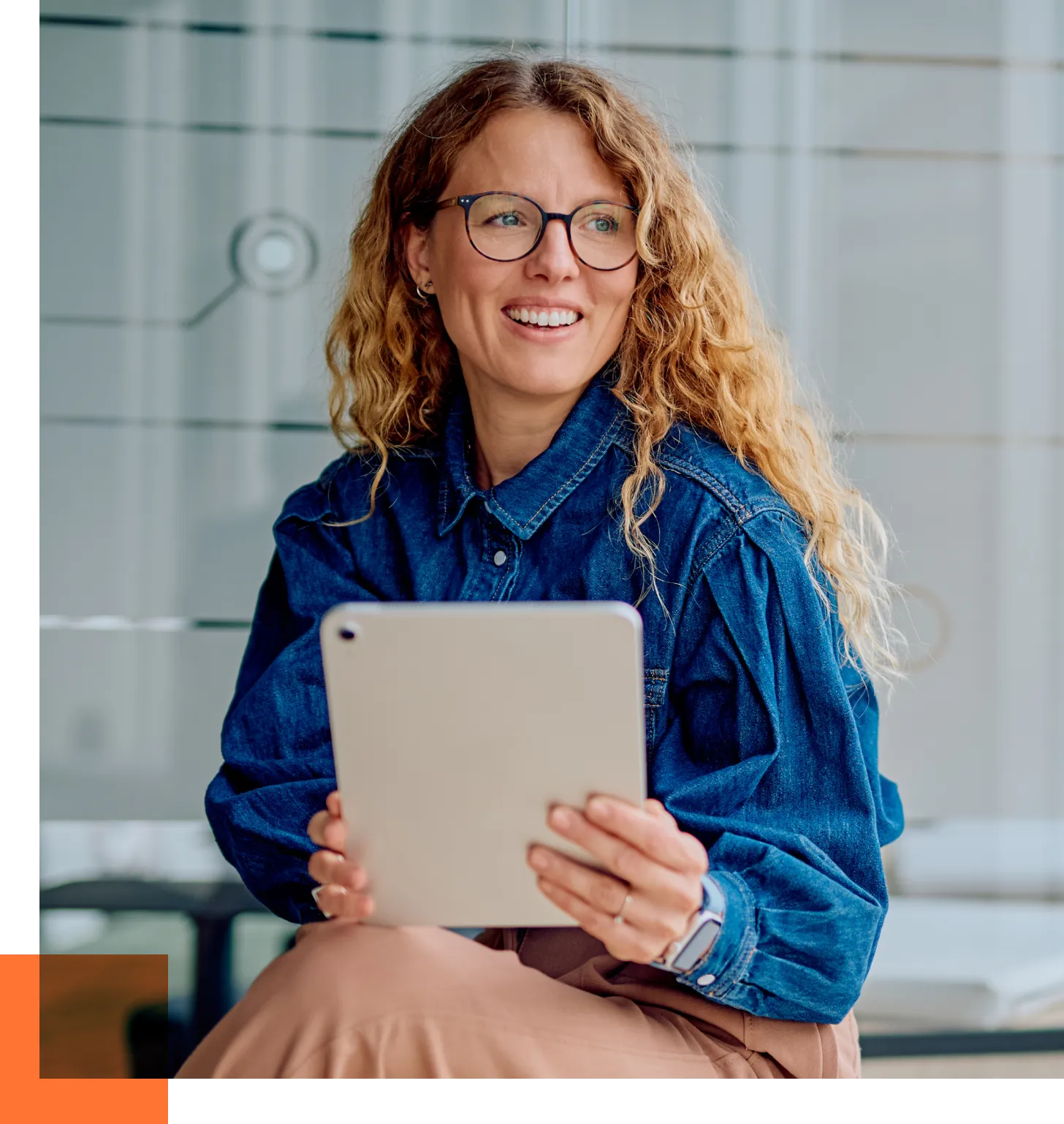 Smiling woman with curly blonde hair and glasses holding a tablet in a modern office setting.