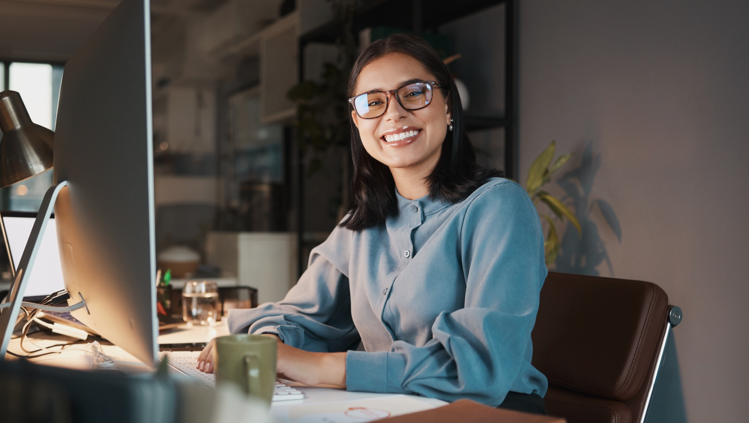 Smiling woman in glasses sits at a desk with a large monitor in a modern office.