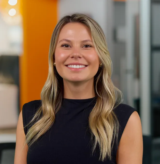 Smiling blonde woman in a black sleeveless top, standing in a bright office with an orange accent wall behind her.