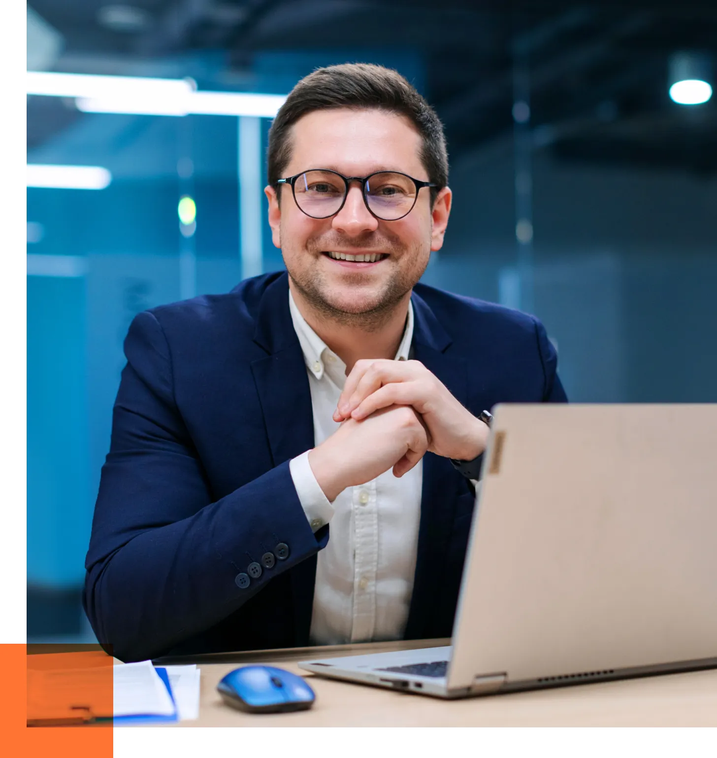Smiling man in a navy suit and glasses sits at a desk with a laptop in a modern office backdrop.