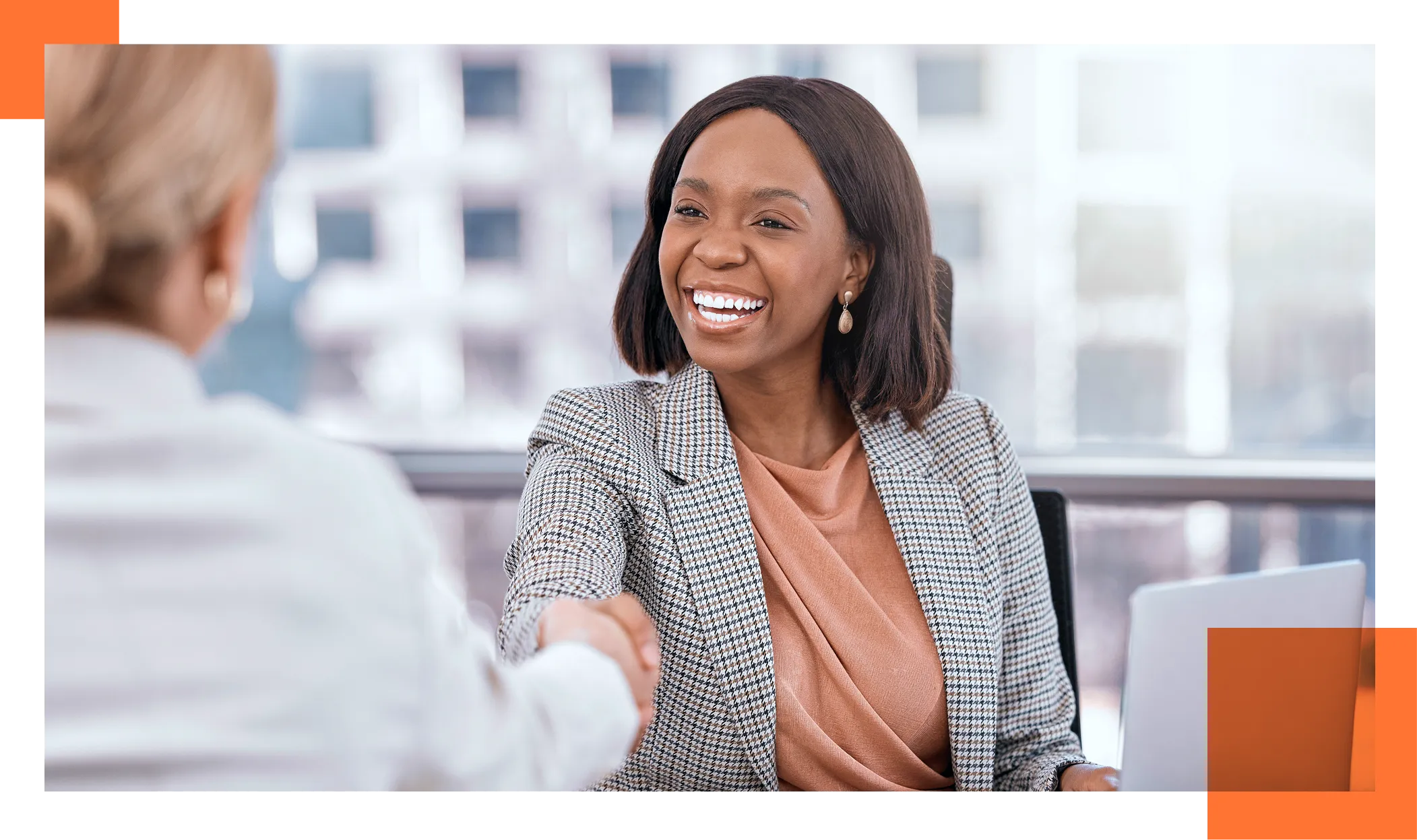 Professional woman in a checkered blazer smiles while shaking hands with a colleague in a bright office setting, indicating a business meeting.