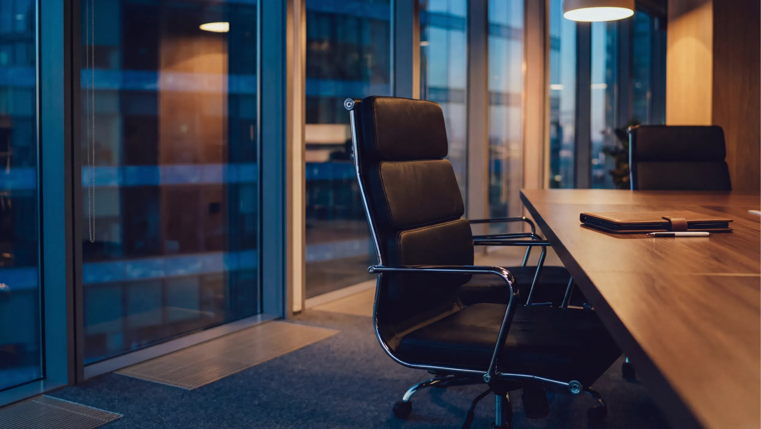 Modern conference room with a black leather chair at a wooden table, notebook and pen nearby Empty office meeting room featuring a cushioned chair, glass walls, and a notebook on a long table Sleek boardroom setup
